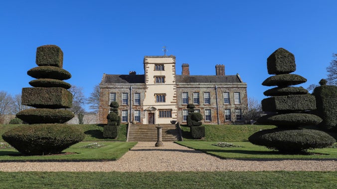 The garden at Canons Ashby, Northamptonshire in February, with neatly trimmed topiary trees and a sundial in the middle of the path, with steps leading up to further gardens and the house beyond